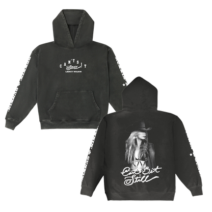 Black hoodie featuring a white graphic print of lainey wilson and the text ’can’t sit still’ on both front and back, with star patterns along the sleeves.
