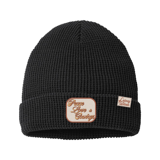 Black waffle-knit beanie featuring a cream-colored patch with ’peace love & cowboys’ embroidered in brown script and a small rectangular tag reading ’lainey wilson.’