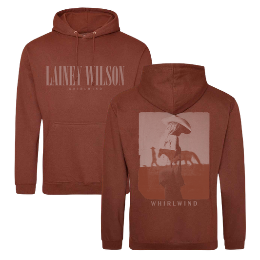 Rust-colored hoodie featuring ’lainey wilson’ and ’whirlwind’ branding with a graphic of a figure in a hat walking beside a horse on the back.