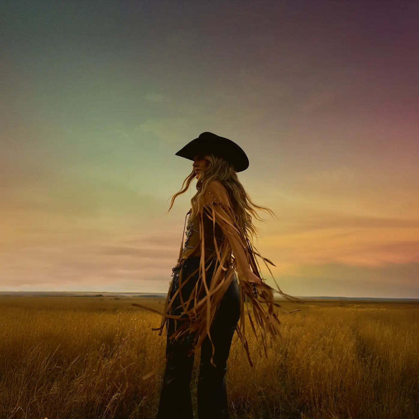 A woman wearing a black cowboy hat with long, flowing blonde hair and a fringed top stands in a golden field at sunset.